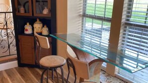 Un comedor con una mesa de vidrio y dos taburetes de bar, un mueble de madera con objetos decorativos, y una ventana con persianas.