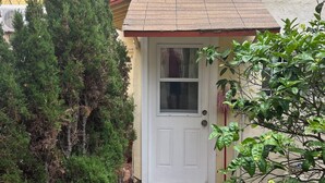 Una piccola casa con una porta bianca, immersa nel verde e un vialetto piastrellato che conduce all'ingresso.