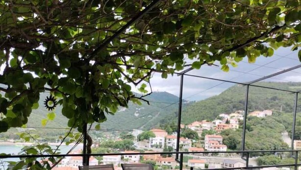 Eine Terrasse mit einem Glastisch und Stühlen, einer Obstschale und einer Flasche, mit Blick auf eine Küstenstadt mit Gebäuden und Hügeln.