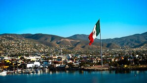 A coastal town with a prominent flag on a flagpole, surrounded by mountains and a marina with boats. - Chic 1-bedroom apartment with ocean views, sun bathing deck, pool in Ensenada. (Ensenada)