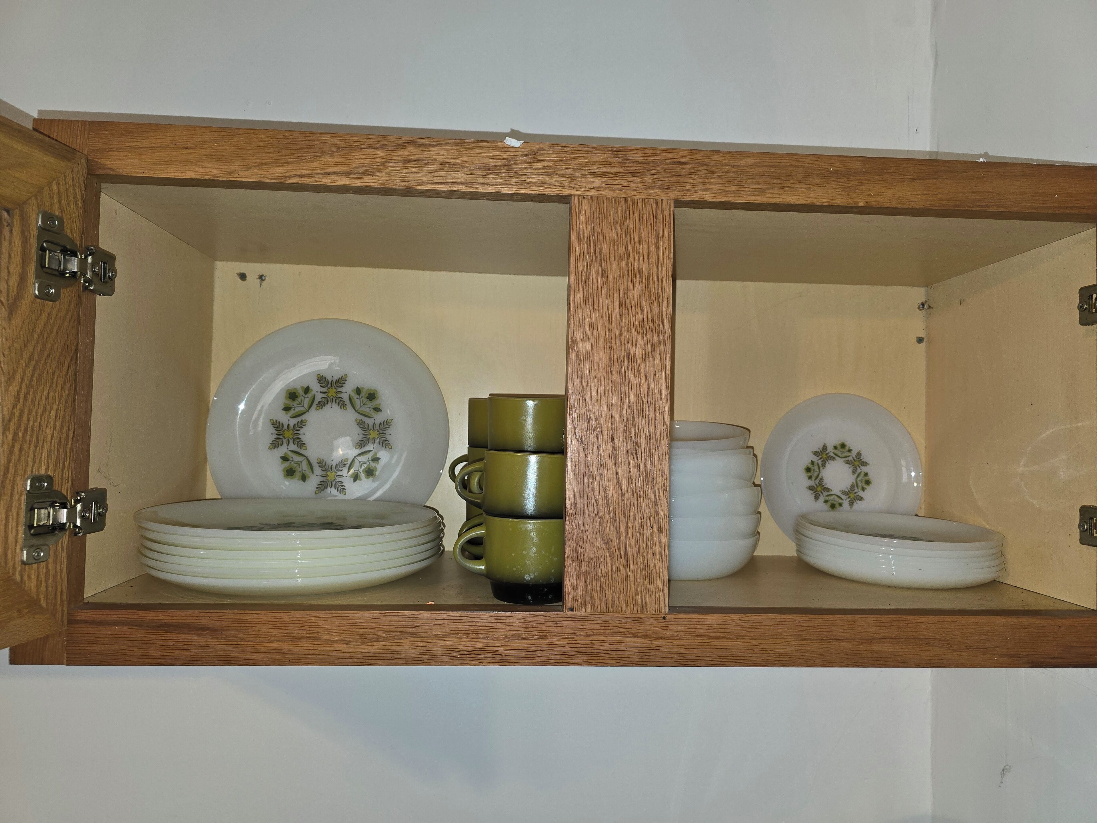 A wooden cabinet with two shelves, each containing white plates with green floral designs, a green cup, and a stack of white plates.