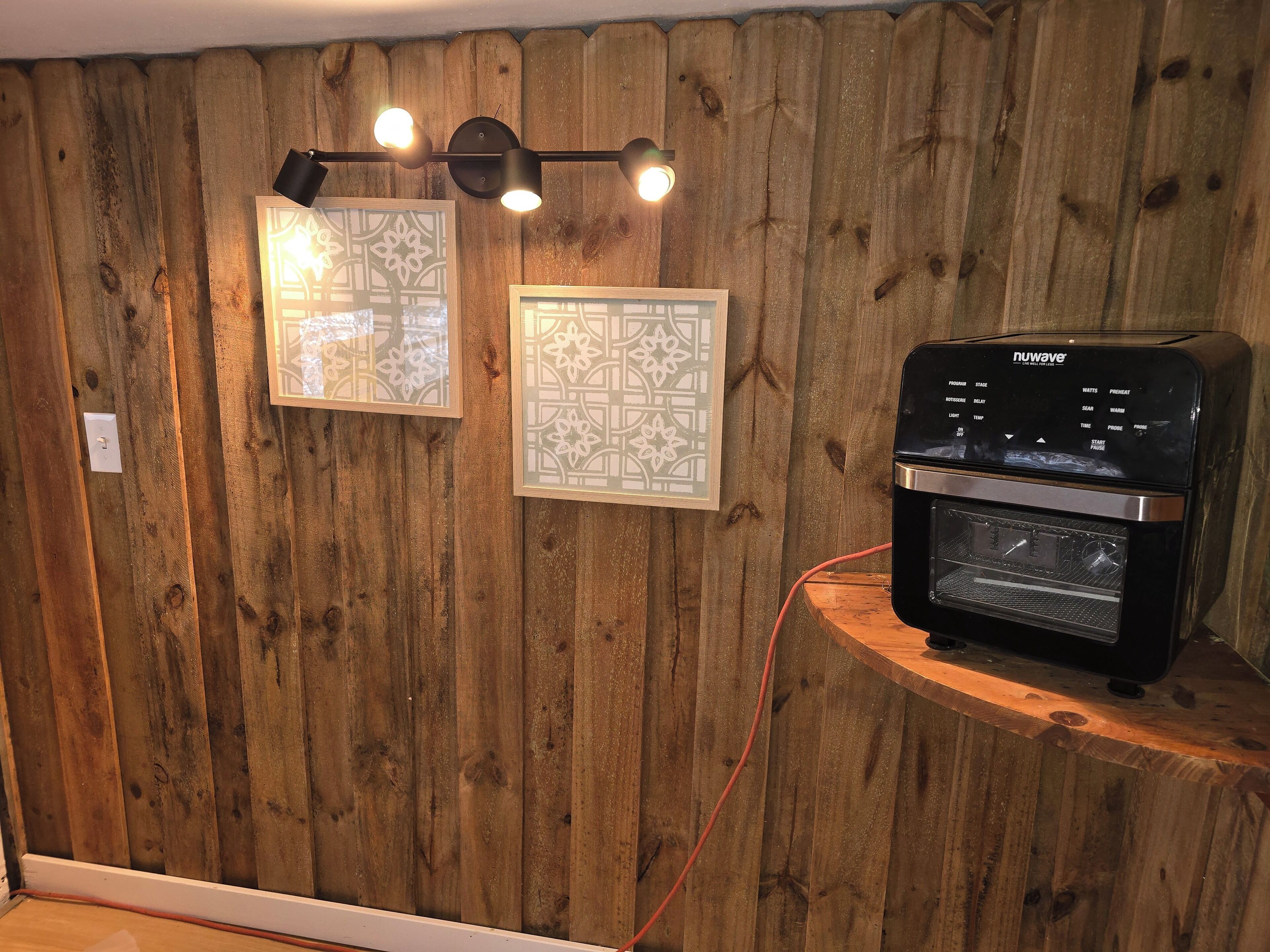 A kitchen with a Nuwave oven, wooden wall paneling, and two framed decorative pieces.