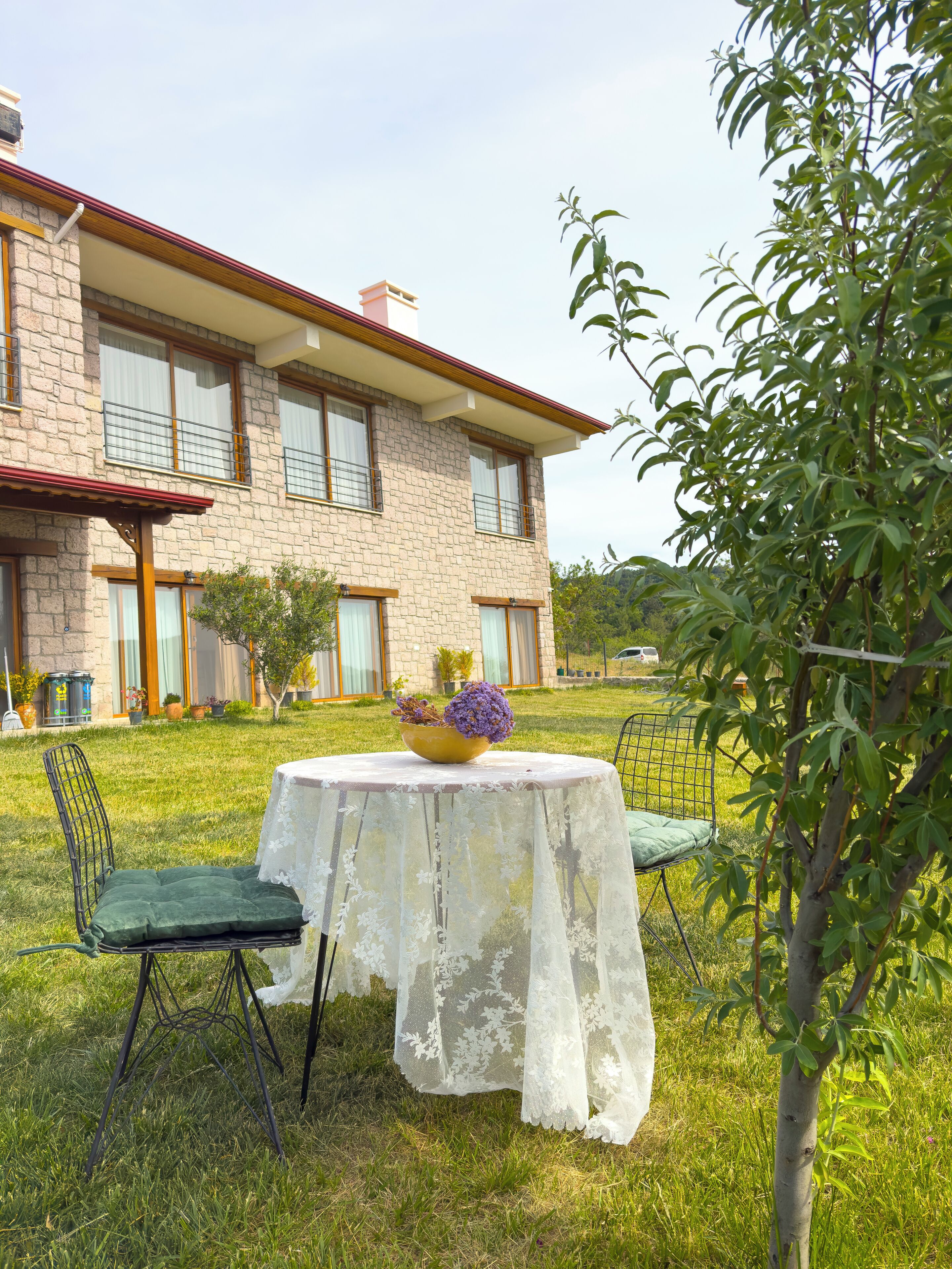 A two-story stone house with a balcony, a round table with a white lace tablecloth, and two chairs.