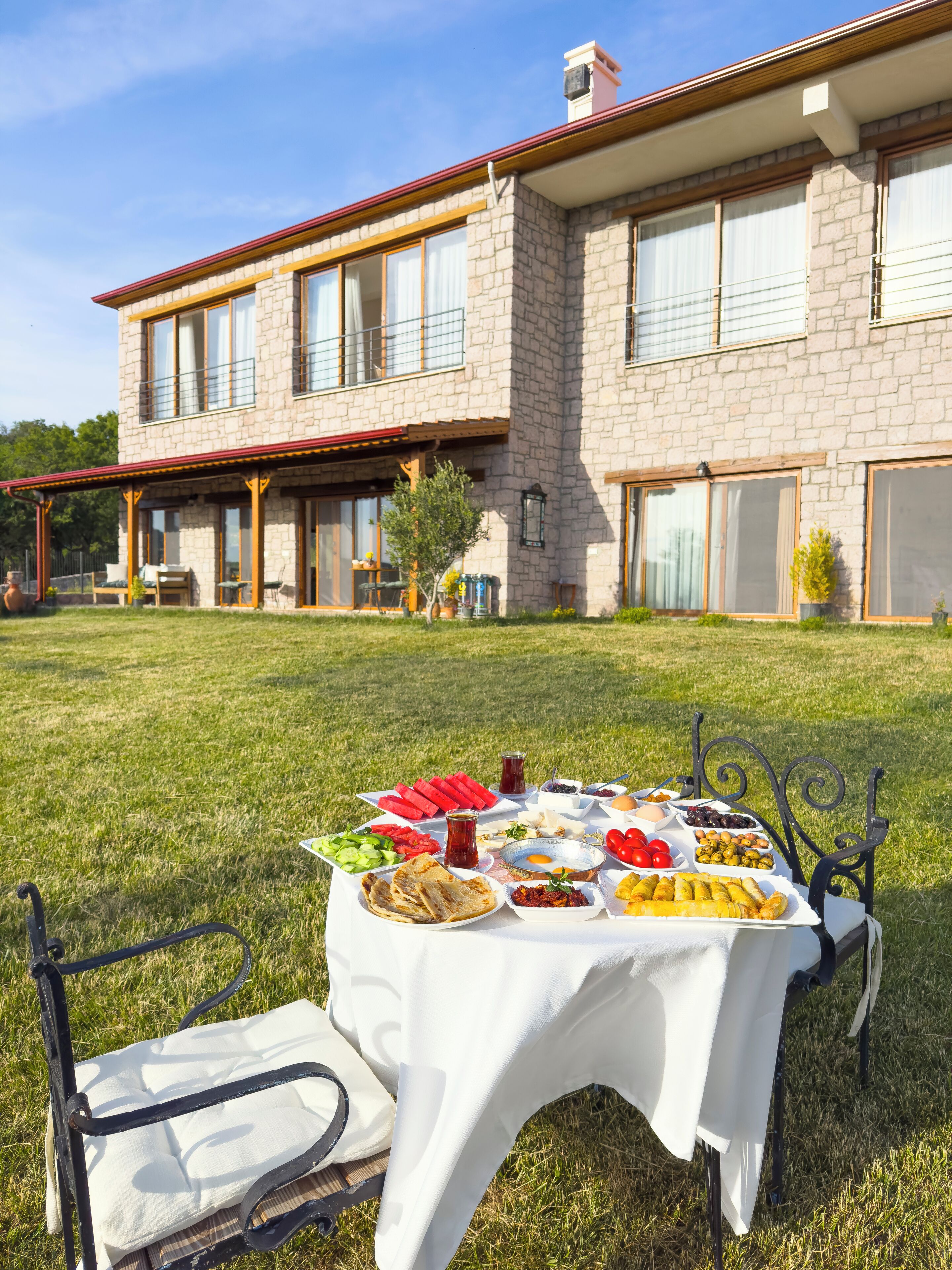 Un espace de restauration en plein air, avec une table dressée pour un repas, entouré d’une maison en pierre avec un toit rouge et de grandes fenêtres.