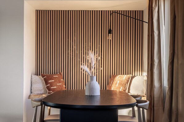 Un comedor con una mesa redonda, dos sillas, un jarrón con plantas secas y una pared con listones de madera verticales.