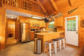 Cocina de cabaña de madera con refrigerador, estufa y lavamanos de acero inoxidable. Hay armarios de madera, una ventana y una puerta con un letrero encima.
