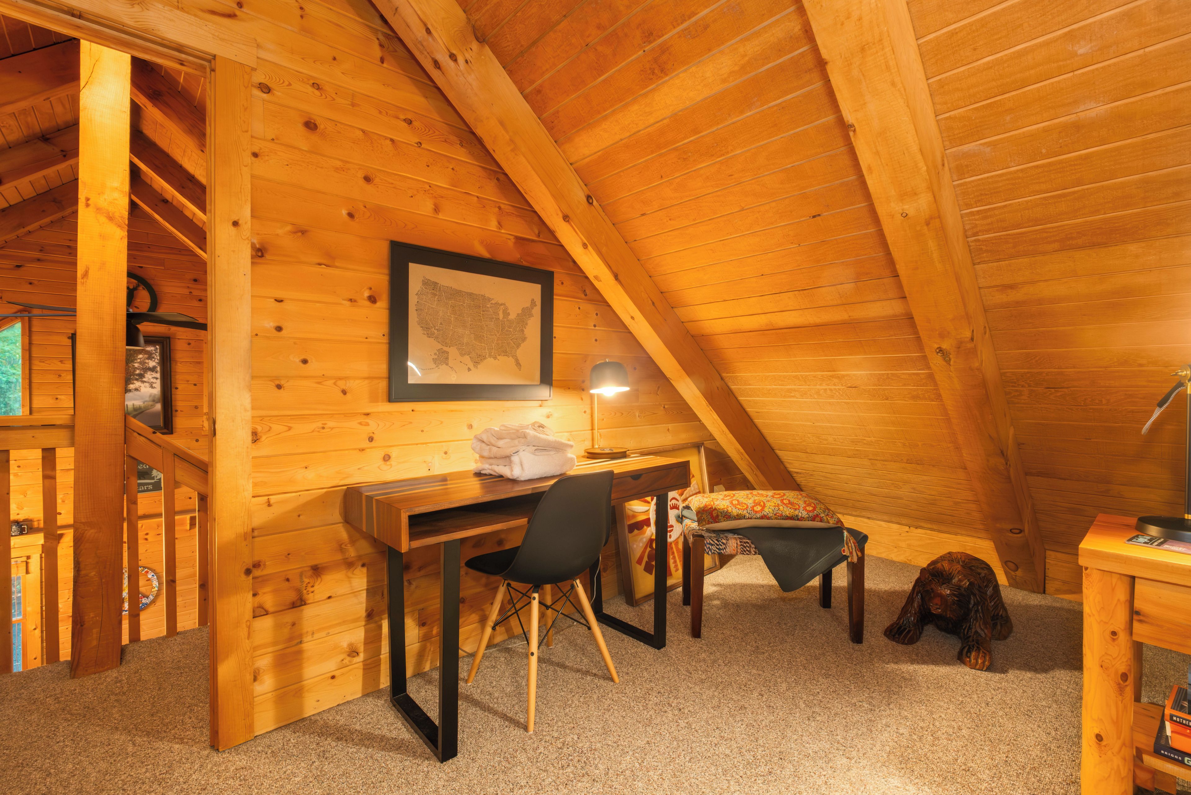 A cozy attic bedroom with a wooden sloped ceiling, a desk with a chair, a bed with a patterned blanket, and a framed map on the wall.