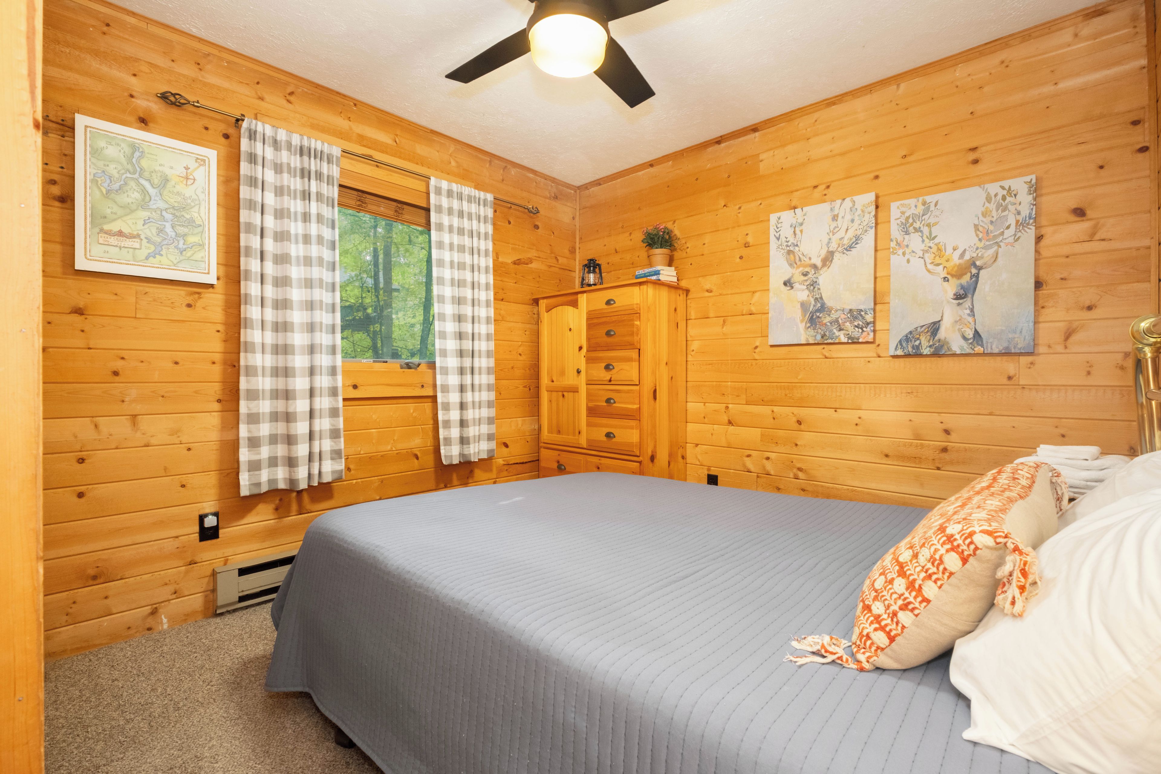 A wooden cabin bedroom with a bed, a dresser, a window with curtains, and two wall-mounted paintings.
