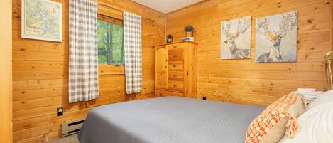 A wooden cabin bedroom with a bed, a dresser, a window with curtains, and two wall-mounted paintings.