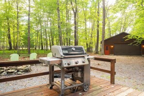 Una parrilla Weber Genesis II portátil sobre una terraza de madera, con vista a un bosque y una cabaña.