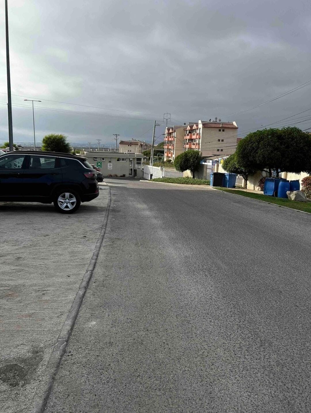 Una carretera con un coche aparcado, un edificio con entrada protegida por una reja y una zona residencial al fondo.