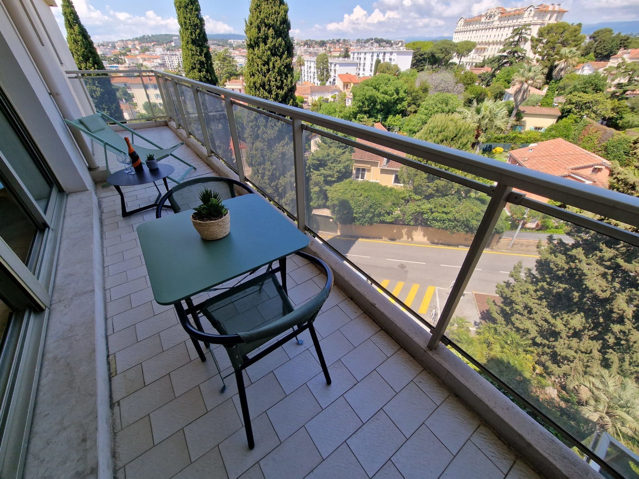 A balcony with a table and chairs, a potted plant, and a view of a cityscape with buildings and trees.