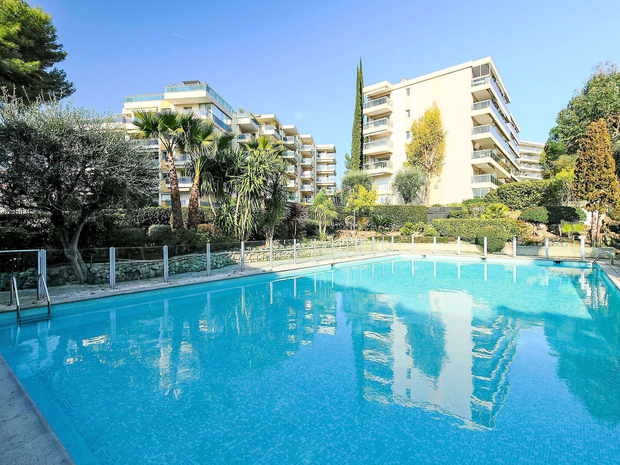 A swimming pool with a clear blue water surface, surrounded by a stone wall and metal railings. In the background, there are multi-story buildings with balconies and a variety of trees and shrubs.