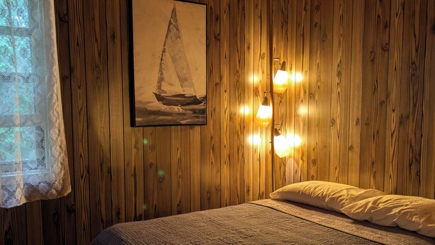 Un dormitorio con paredes de madera, ventilador de techo, un cuadro enmarcado de un velero y luces de lectura fijadas a la pared.