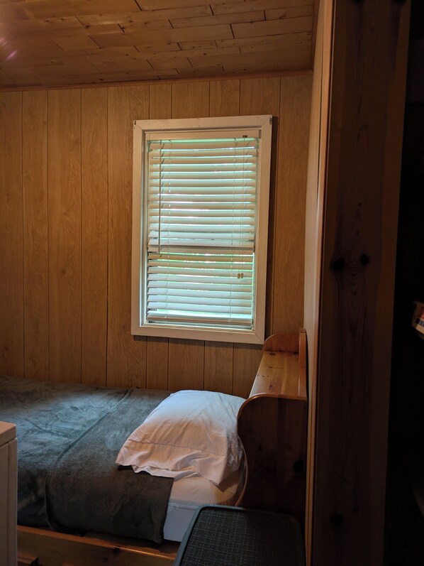 Habitación pequeña con una cama individual, paredes de madera y una ventana con persianas.