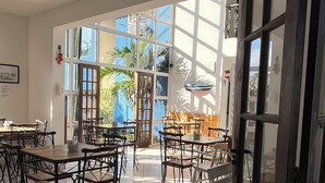 A spacious dining area with multiple tables and chairs, large windows allowing natural light, and a view of palm trees outside. - POUSADA BELUGA (Buzios)