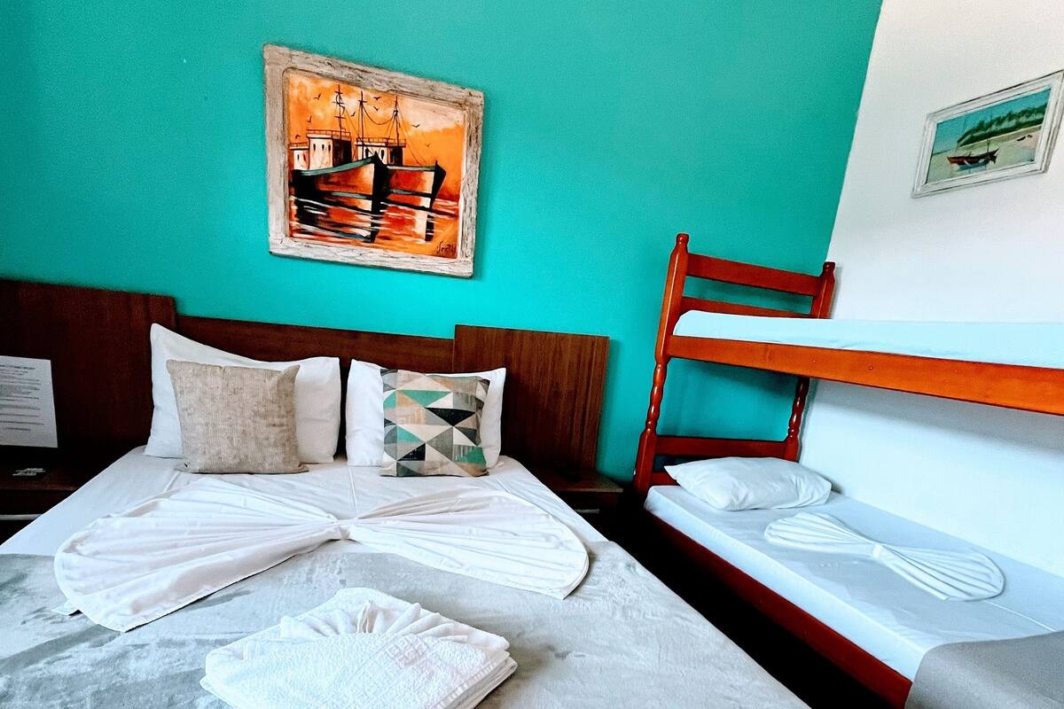 Habitación de hotel con una cama grande, una cama más pequeña y un cabecero de madera. Hay un cuadro en la pared y una ventana con vista al mar.