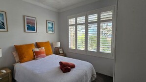 Un dormitorio con una cama, dos almohadas, una mesita de noche, una lámpara y una ventana con persianas.