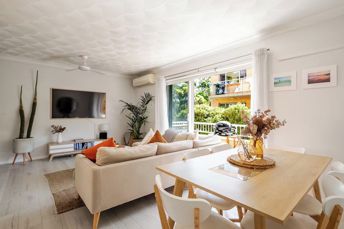 A modern living room with a sofa, dining table, and chairs. There is a large window with a view of greenery and a balcony.