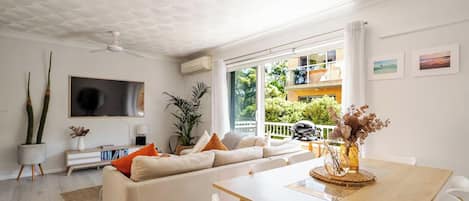 A modern living room with a sofa, dining table, and chairs. There is a large window with a view of greenery and a balcony.