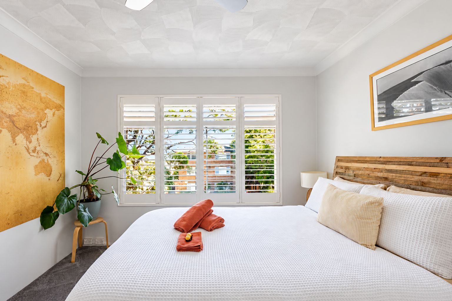 A hotel bedroom with a large bed, a potted plant, and a view through a window with blinds.
