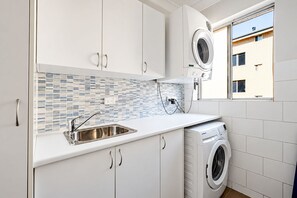 Ett modernt kök med vita skåp, en diskho i rostfritt stål, kakel på väggen bakom diskbänken och en inbyggd tvättmaskin.