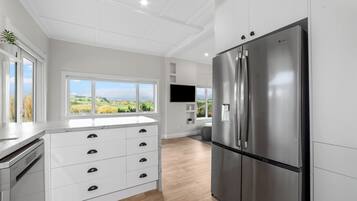 A modern kitchen with a stainless steel refrigerator, white cabinetry, and a large window offering a view of the countryside.