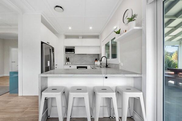 A modern kitchen with white cabinetry, a marble countertop, and a stainless steel refrigerator. There are four white stools under the counter.