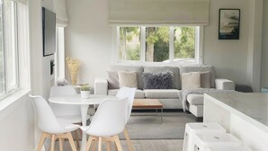 Un salon moderne et bien éclairé, avec un canapé gris, des chaises blanches, une petite table et un coin cuisine doté de tabourets blancs.