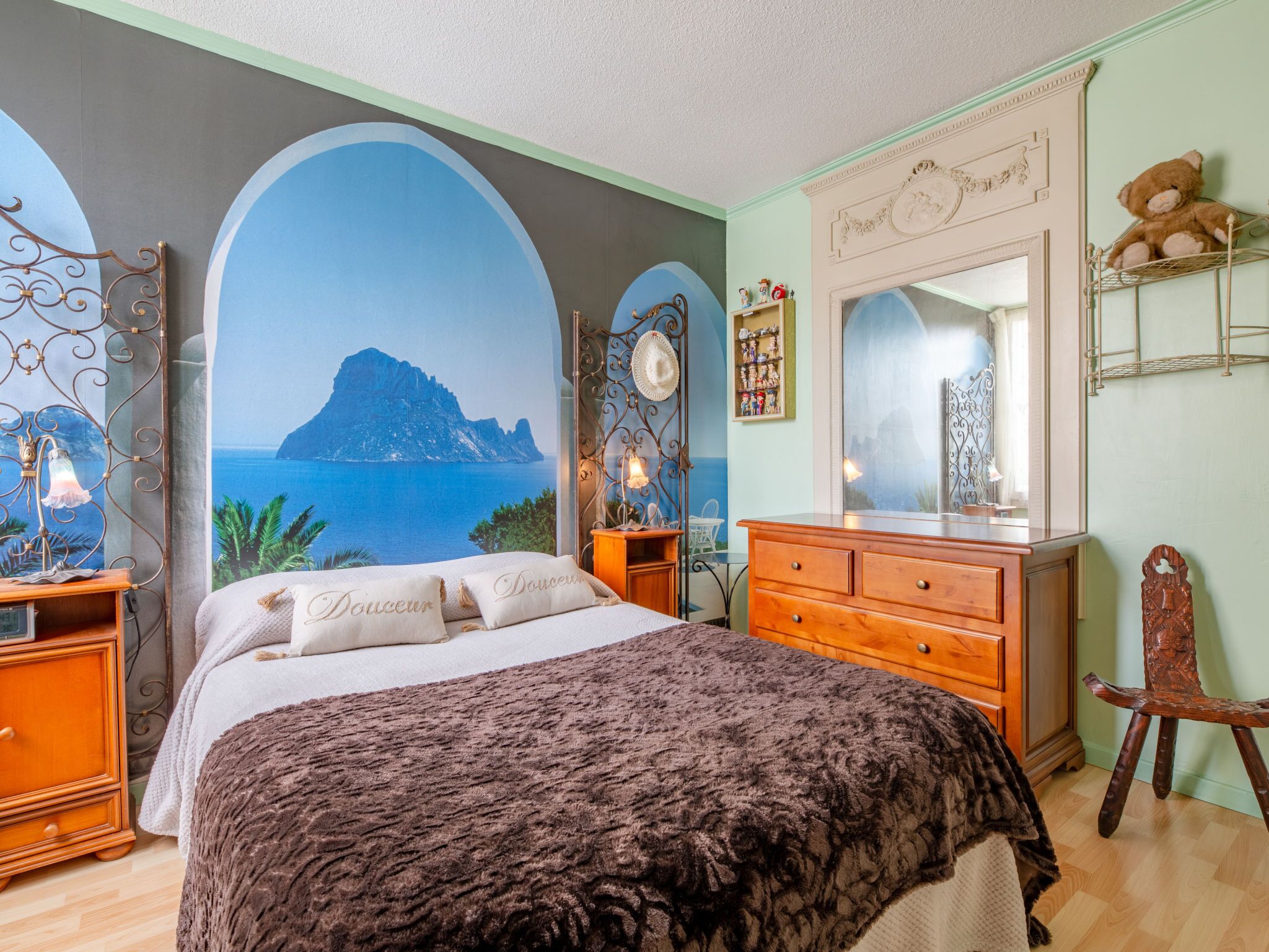 Een slaapkamer met een bed, een dressoir, een stoel en een groot muurschildering van een kustlandschap.
