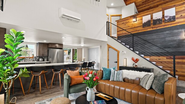 A modern kitchen with a central island, a dining area with bar stools, a living room with a leather sofa, and a staircase with a wooden railing.