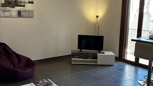 A modern living room with a flat-screen TV, a white coffee table, a bean bag, and a large window providing natural light.