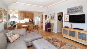 Una sala de estar con un sofá seccional gris, un comedor con mesa y sillas de madera, una cocina con gabinetes de madera, un televisor de pantalla plana montado en la pared y un tapete estampado.