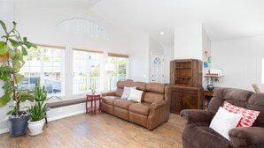 A spacious living room with a brown leather sofa, a red armchair, and a wooden cabinet. There are two potted plants and a small red stool. - 364 Harbor View (Pismo Beach)
