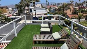 Een terras op het dak met kunstgras, twee houten lounge stoelen en een witte balustrade.