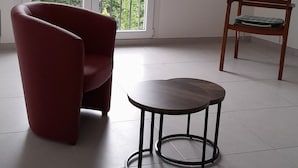 Een kamer met een rode stoel, een klein rond tafeltje en een houten stoel bij een groot raam met uitzicht op groen.
