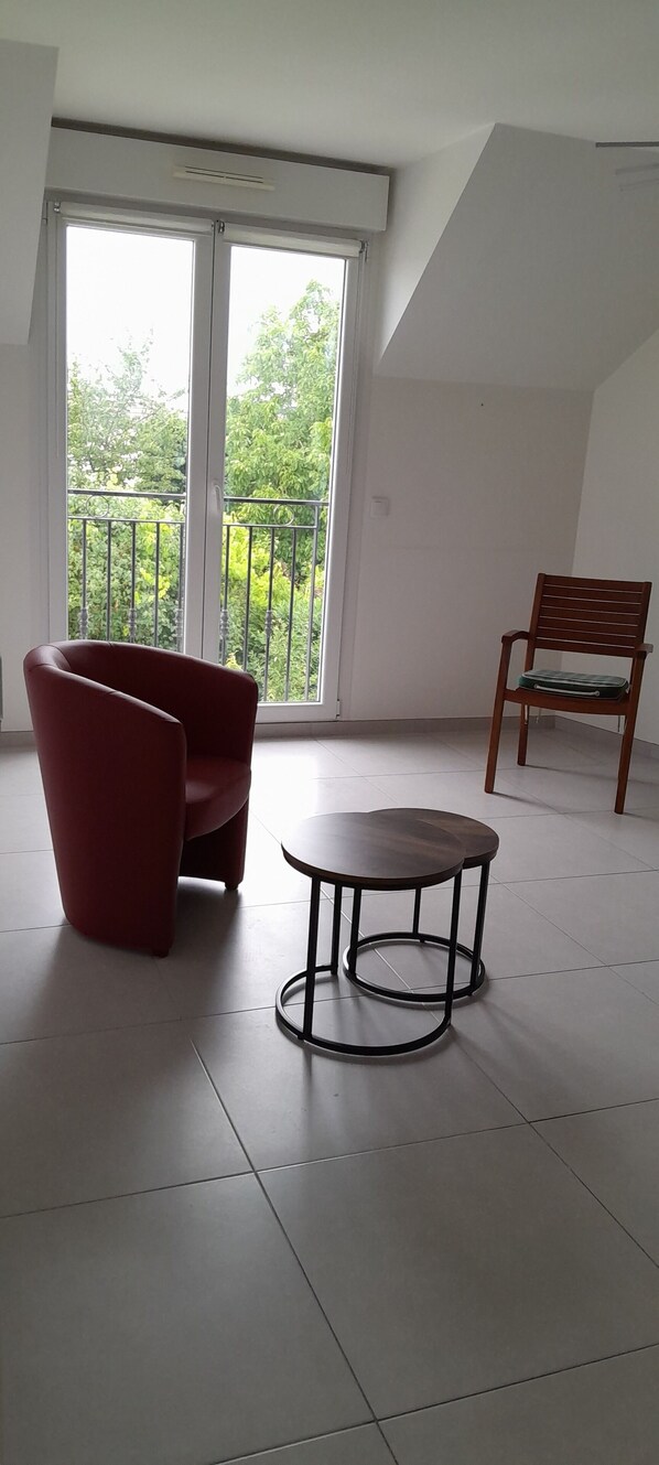 A room with a red chair, a small round table, and a wooden chair near a large window with a view of greenery.