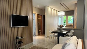 A modern living room with a wooden sofa, a flat-screen TV mounted on the wall, and a dining area with a table and chairs.