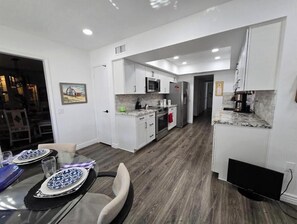 A modern kitchen with white cabinets, a microwave, and a refrigerator. There is a dining area with a glass table and chairs. - Welcome to beautiful Desert Glen Inn located in the "Arizona Valley of the Sun" (Sun City West)