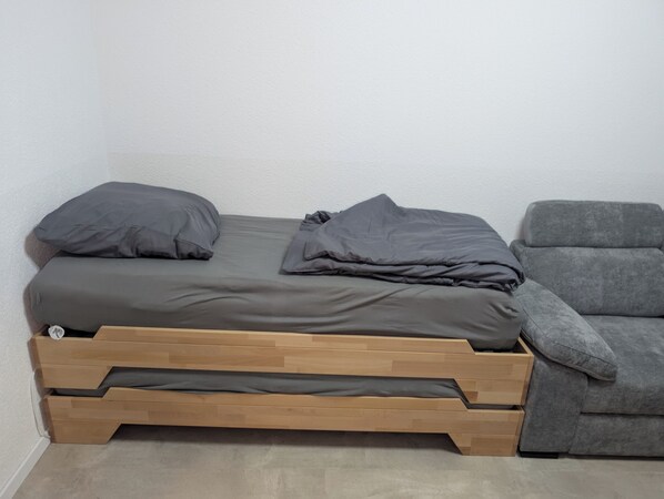 A compact bed with a wooden frame and grey bedding, adjacent to a grey armchair in a room with plain white walls.