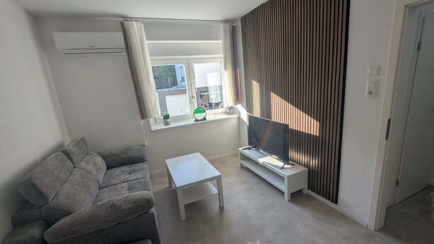 A modern living room with a grey sofa, a small white table, and a flat-screen TV on a white stand. - Small city apartment Remscheid (Remscheid)
