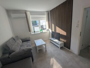 A modern living room with a grey sofa, a small white table, and a flat-screen TV on a white stand. - Small city apartment Remscheid (Remscheid)