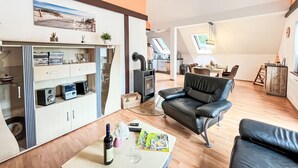 A modern living room with a black leather sofa, a glass coffee table, and a bookshelf. There is a kitchen area with a stove and a dining table in the background.