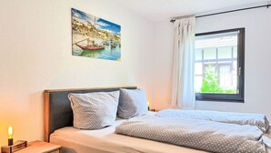 A bedroom with a bed, a nightstand, a painting of a coastal scene, a window with curtains, and a view of greenery outside.