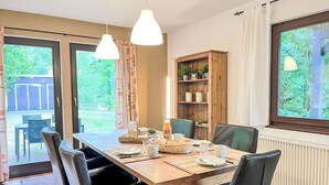 A dining area with a wooden table, black chairs, and hanging pendant lights. There is a window with a view of greenery outside.