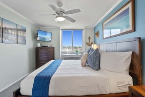 A bedroom with a bed, a ceiling fan, a TV, a dresser, and a window with a view of the beach.