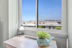Un escritorio con una lámpara, un libro y una planta junto a una ventana que da a una playa y edificios.