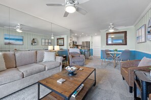 Ein modernes Wohnzimmer mit Sofa, Couchtisch und Sesseln, ein Küchenbereich mit Geräten sowie ein Esstisch mit Stühlen.