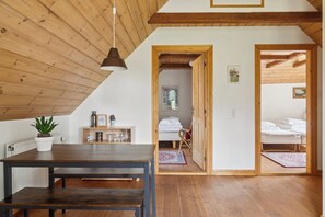 Ein Zimmer mit Holzbalkendecke, einem Esstisch mit Bänken, einer Kübelpflanze und einem Bett mit weißer Daunendecke.