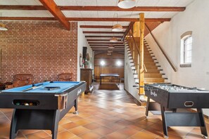 Une table de billard, une table de baby-foot et un escalier dans une pièce dont un mur est en briques et dont les poutres sont en bois.